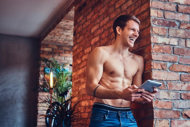 Un uomo sexy a torso nudo tatuato con il touchpad si appoggia al muro di mattoni. Sorridere e distogliere lo sguardo.