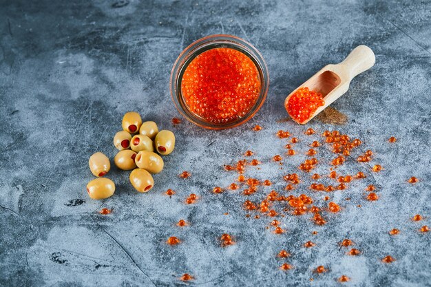 Un vasetto di caviale rosso e olive verdi su sfondo marmo.