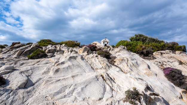 Un vecchio e piccolo santuario situato sulle rocce vicino alla costa del Mar Egeo, cespugli intorno, cielo nuvoloso, Grecia