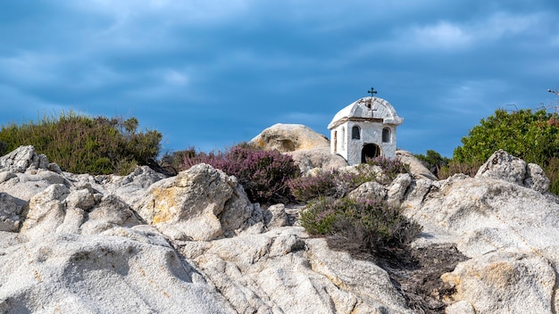 Un vecchio e piccolo santuario situato sulle rocce vicino alla costa del Mar Egeo, cespugli intorno, cielo nuvoloso, Grecia