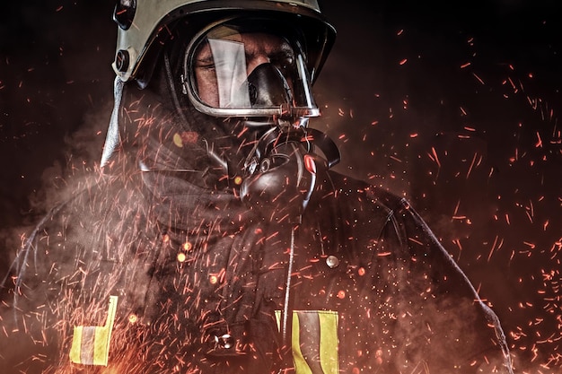 Un vigile del fuoco professionista vestito in uniforme e una maschera di ossigeno in piedi in scintille di fuoco e fumo su uno sfondo scuro.