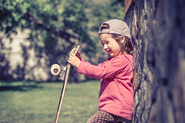 Una bambina alla moda tiene in mano uno skateboard e gioca fuori, le belle emozioni di un bambino.
