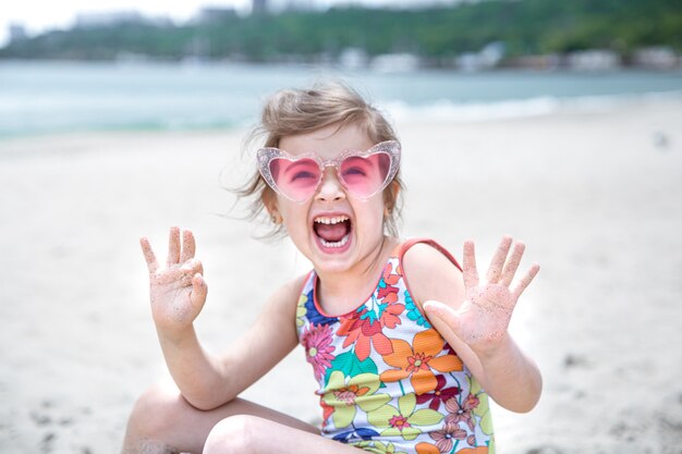 Una bambina carina con gli occhiali sta giocando nella sabbia sulla spiaggia in riva al mare.