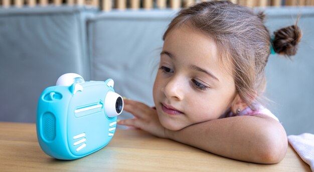 Una bambina carina guarda la macchina fotografica giocattolo blu dei suoi figli per la stampa di foto istantanea.