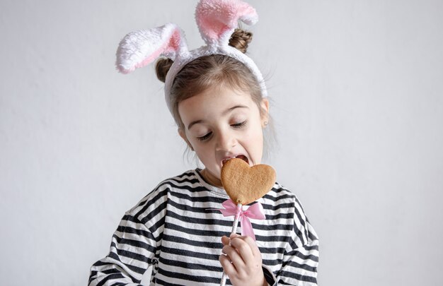 Una bambina carina sta mordendo un pan di zenzero pasquale su un bastone e con le orecchie da coniglio decorative sulla testa.