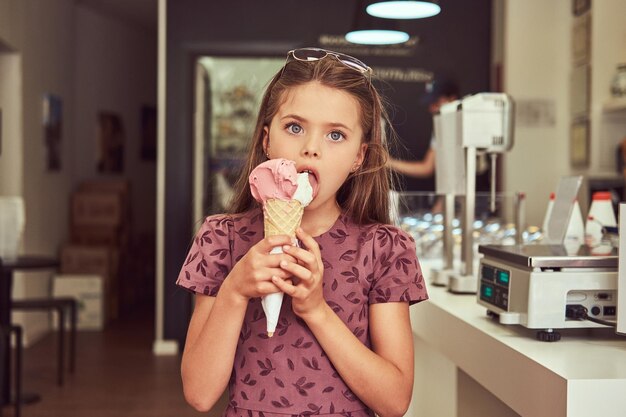 Una bambina di bellezza in un vestito alla moda che mangia fragola, in piedi in una gelateria.