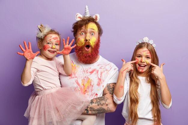 Una bambina piccola mostra le mani sporche di pitture a guazzo colorate, la sorella felice ha macchie di acquerello sul viso, il padre sbalordito fissa, si diverte prima che la madre torni dal lavoro.