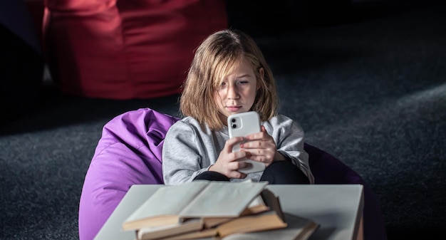 Una bambina si sdraia su un pouf e usa uno smartphone invece di leggere un libro