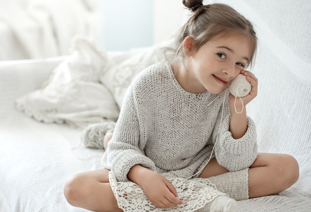 Una bambina si siede sul divano con un gomitolo di filo e impara a lavorare a maglia.