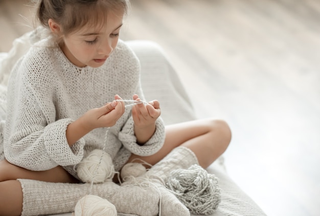 Una bambina si siede sul divano con un gomitolo di filo e impara a lavorare a maglia.