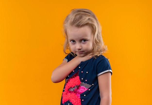 Una bella bambina con i capelli biondi che indossa la camicia blu navy che tiene la sua gola su una parete arancione