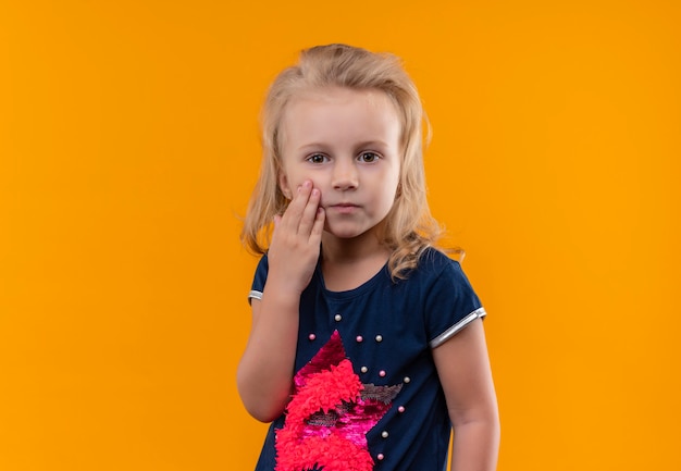 Una bella bambina con i capelli biondi che indossa la camicia blu navy che tocca il suo fronte su una parete arancione