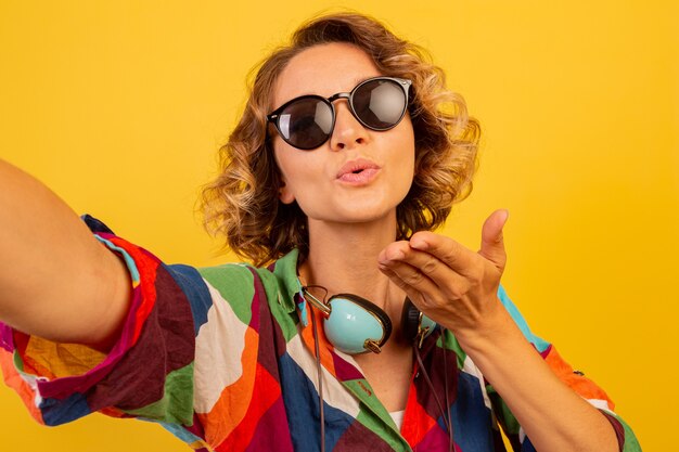 Una bella donna carina con gli auricolari manda un bacio alla telecamera e fa un autoritratto sul muro giallo