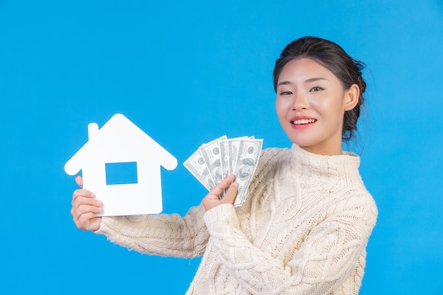 Una bella donna che indossa un nuovo tappeto bianco a maniche lunghe che detiene il simbolo della casa e banconote da un dollaro su un blu. Trading.