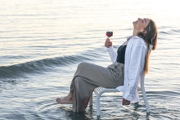 Una bella donna con un bicchiere di vino in riva al mare si siede su una sedia