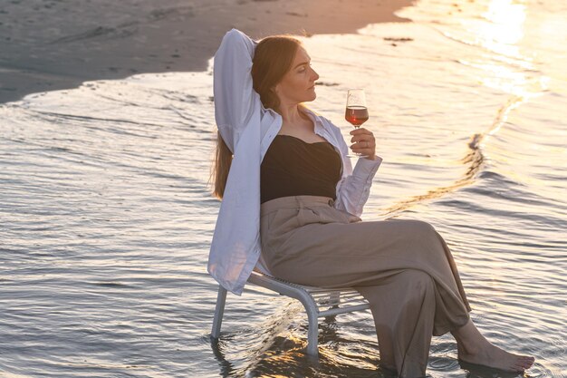 Una bella donna con un bicchiere di vino in riva al mare si siede su una sedia