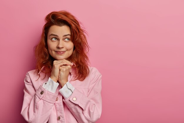 Una bella donna dai capelli rossi tiene le mani sotto il mento, ha un aspetto piacevole, sogna che sia accaduto qualcosa di molto buono, indossa abiti alla moda