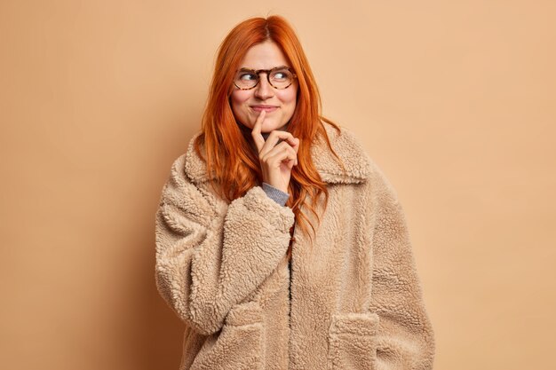 Una bella donna sognante con i capelli rossi guarda da parte pensieroso indossa un cappotto invernale marrone.