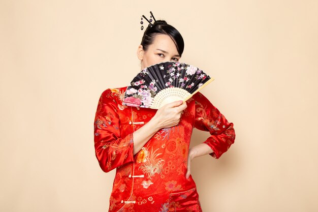 Una bella geisha giapponese di vista frontale in vestito giapponese rosso tradizionale con i bastoncini di capelli che posano tenendo il ventaglio pieghevole elegante sulla cerimonia crema giapponese del fondo