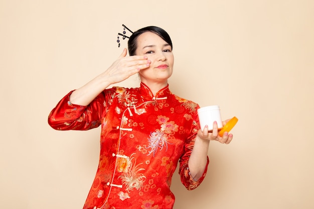 Una bella geisha giapponese di vista frontale in vestito giapponese rosso tradizionale con i bastoncini di capelli che posano usando la crema può odorare sulla cerimonia del fondo crema Giappone