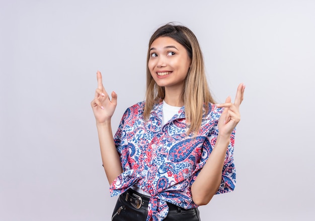 Una bella giovane donna che indossa la camicia stampata paisley che attraversa il dito come gesto di buona fortuna o speranza su un muro bianco