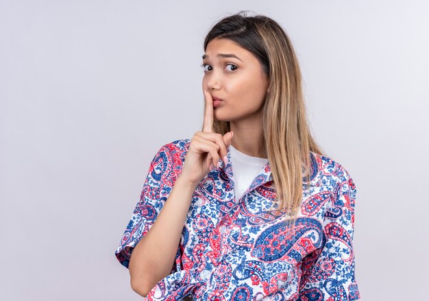 Una bella giovane donna che indossa la camicia stampata paisley che mostra il gesto di shh con il dito mentre guarda su un muro bianco