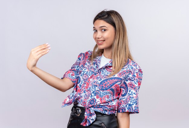Una bella giovane donna che indossa una camicia stampata paisley alzando la mano mentre guarda su un muro bianco