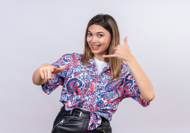 Una bella giovane donna che indossa una camicia stampata paisley che mostra chiamami gesto mentre punta su un muro bianco