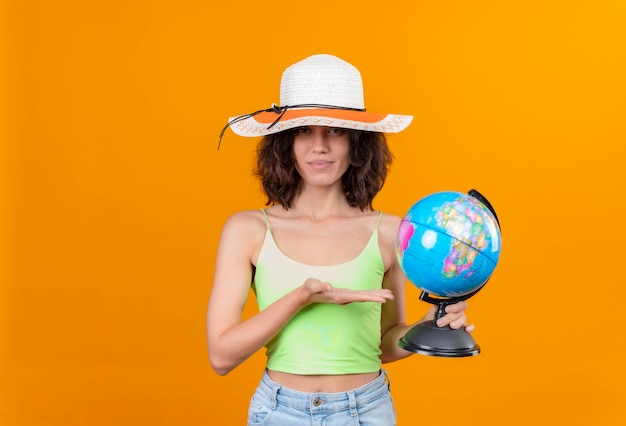 Una bella giovane donna con i capelli corti in top corto verde che indossa un cappello da sole sorridendo e mostrando un globo