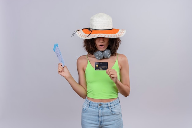 Una bella giovane donna con i capelli corti in verde crop top in cuffia che indossa cappello da sole in possesso di biglietti aerei guardando la carta di credito su uno sfondo bianco