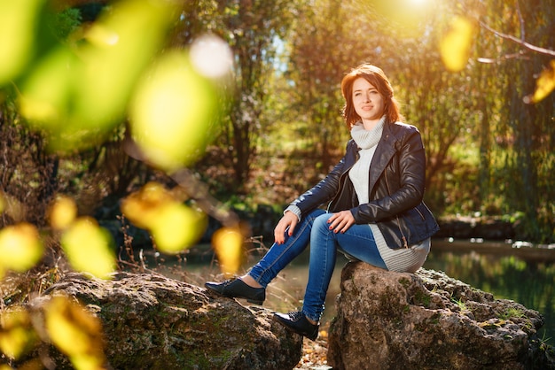 Una bella giovane donna è seduta su una roccia vicino a uno stagno in un parco autunnale al sole