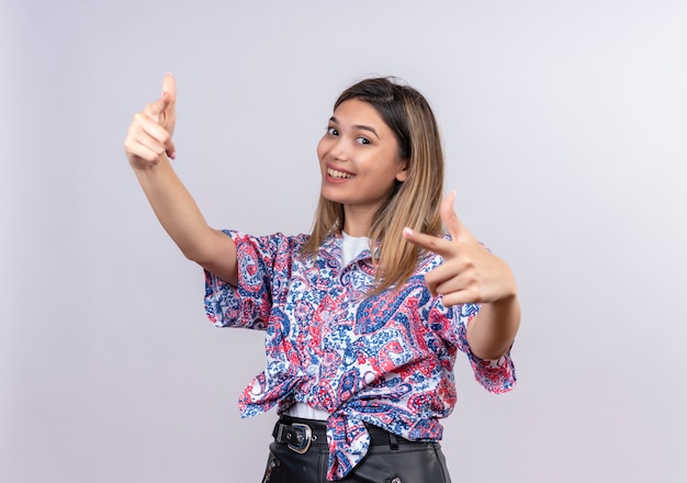 Una bella giovane donna felice che indossa la camicia stampata paisley che punta con il dito indice mentre guarda su un muro bianco