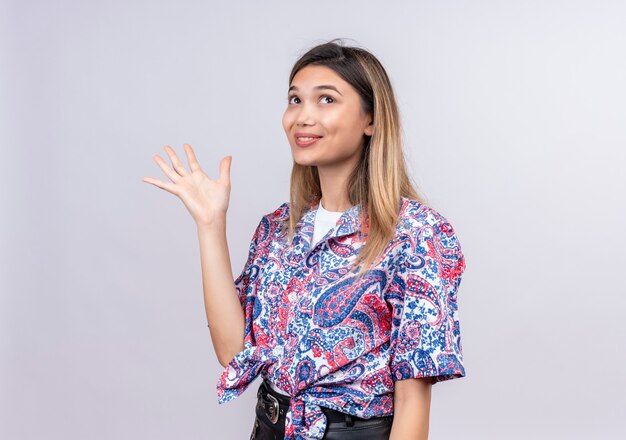Una bella giovane donna felice che indossa la camicia stampata paisley che solleva la mano mentre guarda verso l'alto su un muro bianco