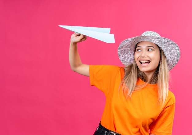 Una bella giovane donna felice e gioiosa in una maglietta arancione che indossa cappellino da sole sorridente mentre vola aeroplano di carta su una parete rosa