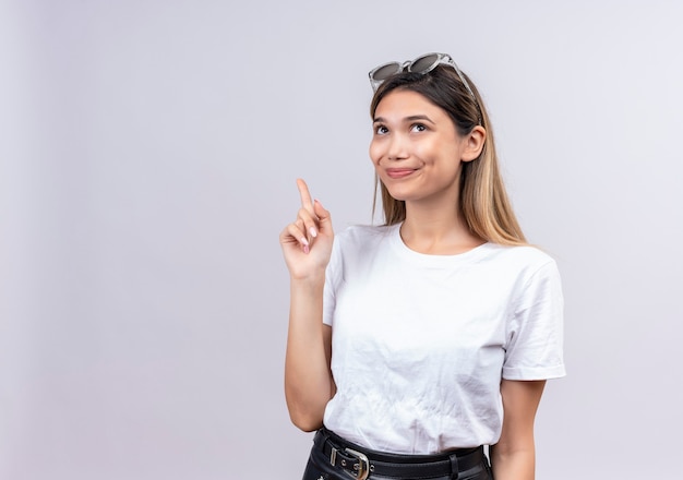 Una bella giovane donna felice in maglietta bianca che indossa occhiali da sole sulla sua testa alzando il dito indice verso l'alto pur avendo un suggerimento su un muro bianco