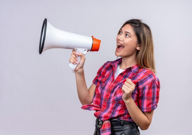 Una bella giovane donna in camicia a quadri parlando tramite il megafono su un muro bianco