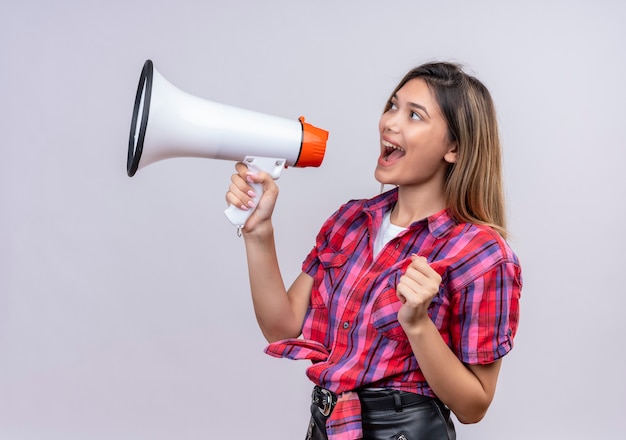 Una bella giovane donna in camicia a quadri parlando tramite il megafono su un muro bianco