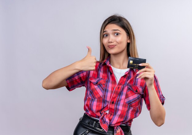 Una bella giovane donna in camicia a quadri sorridente mentre mostra la carta di credito con il pollice in alto su un muro bianco