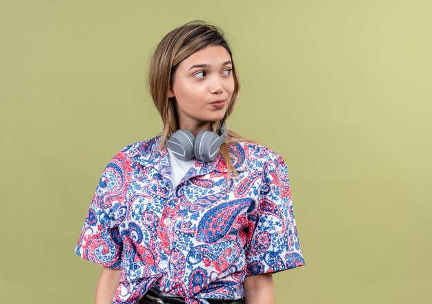 Una bella giovane donna in camicia stampata cachemire che indossa le cuffie pensando e guardando di lato su una parete verde