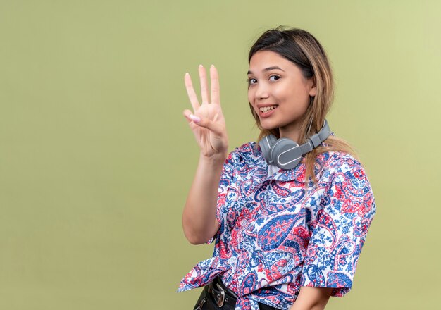 Una bella giovane donna in camicia stampata paisley che indossa le cuffie sorridendo e mostrando tre numeri su una parete verde