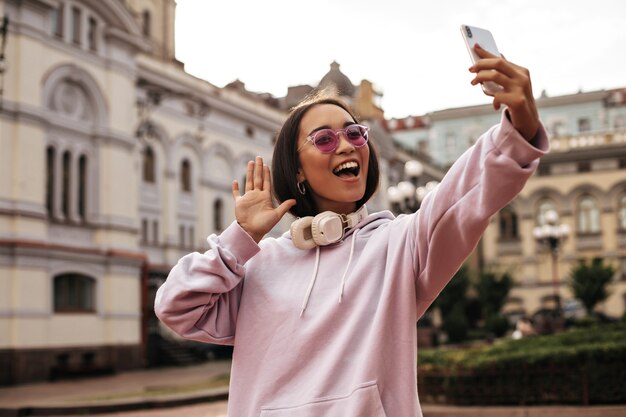 Una bella giovane donna in felpa rosa con cappuccio e occhiali da sole alla moda si fa selfie, tiene il telefono e si mette in posa con le cuffie all'esterno