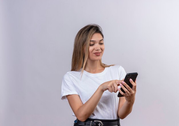 Una bella giovane donna in maglietta bianca sorridente mentre si tocca lo schermo del telefono cellulare su un muro bianco