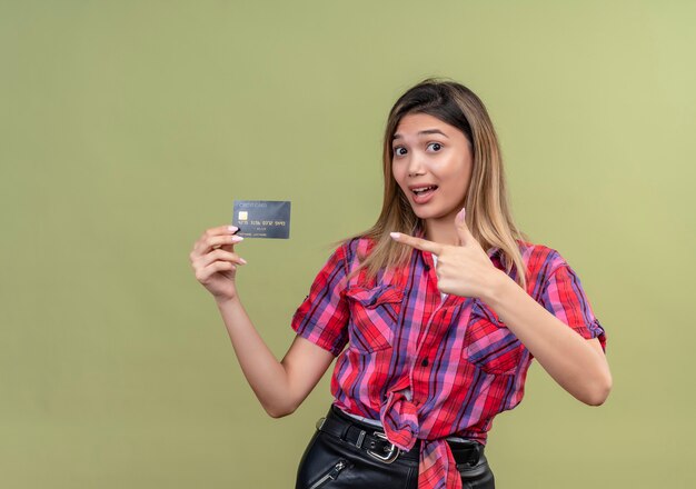 Una bella giovane donna in una camicia a quadri che mostra la carta di credito e indica con il dito indice