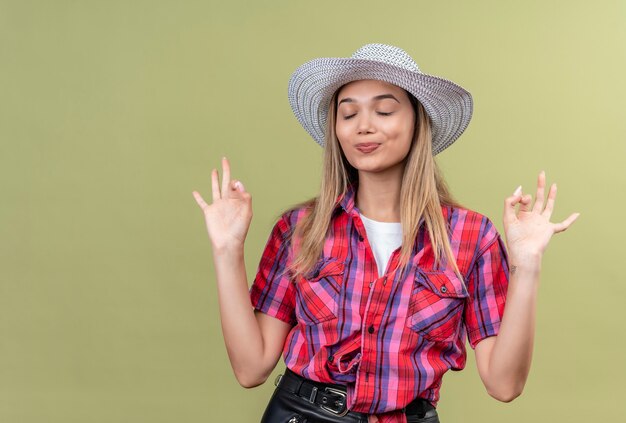 Una bella giovane donna in una camicia a quadri in cappello che chiude gli occhi mentre mostra il gesto giusto su un muro verde