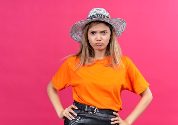 Una bella giovane donna scontenta in una maglietta arancione che indossa un cappello da sole tenendo le mani sulla vita mentre guarda un muro rosa