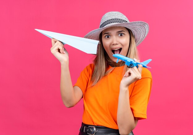 Una bella giovane donna sorpresa in una maglietta arancione che indossa un cappellino da sole che vola in aeroplano di carta mentre tiene un aereo giocattolo blu su una parete rosa