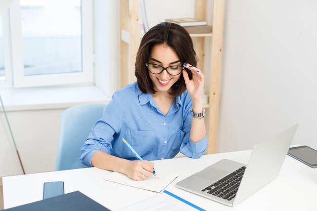 Una bella ragazza bruna con una camicia blu sta lavorando al tavolo in ufficio. Sta scrivendo sul taccuino con un sorriso.