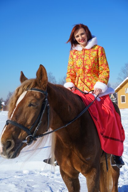 Una bella ragazza in una calda giacca giallo-rossa cavalca un cavallo in una soleggiata giornata gelida invernale. Pratica sport equestri nella stagione invernale