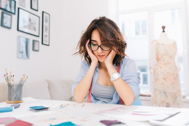 Una bella ragazza mora è seduta al tavolo nello studio del laboratorio. La ragazza in camicia blu è impegnata a guardare gli schizzi nell'album.