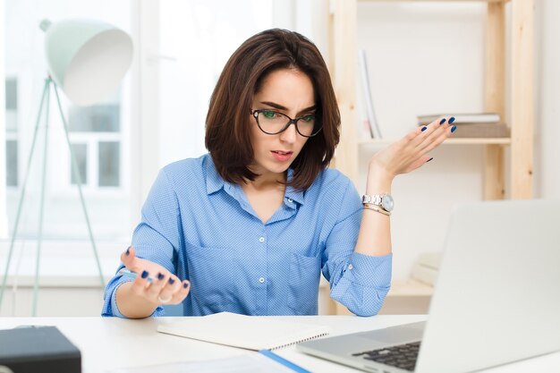 Una bella ragazza mora in camicia blu è seduta al tavolo in ufficio. È sorpresa e persa dal lavoro.